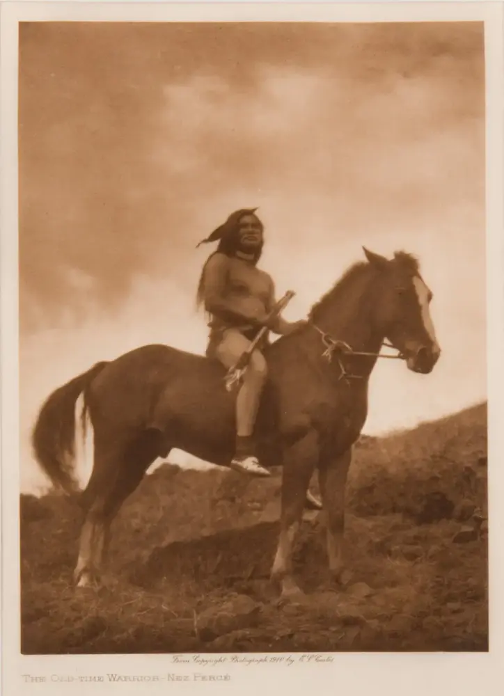 Sepia-toned photograph of an old-time Native American warrior bare-chested, riding a horse across a grassy hillside, holding a spear.