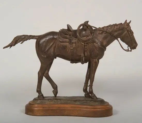 Bronze statue of a saddled horse with a coiled lariat on its back, mounted on a wooden pedestal.