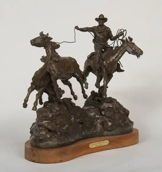 Bronze sculpture of a cowboy on a rearing horse roping a steer, set on a rocky base with a wooden plaque baseplate
