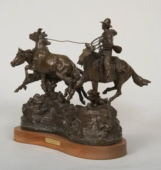 Bronze sculpture of a cowboy on a rearing horse roping a steer, with another horse nearby, mounted on a rugged rock base and wooden plinth.