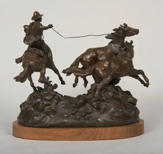 Bronze sculpture of a cowboy on a rearing horse lassoing a bucking horse on a rocky base, mounted on a wooden pedestal.