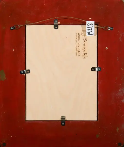 Back of a wooden picture frame mounted on a red wall, showing a pale plywood panel, hanging wire, and metal brackets with a small tag.