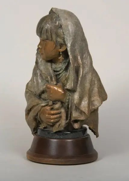 Stone or bronze statue of a seated woman in a hooded cloak, hands clasped near chest, wearing a cross earring, on a wooden pedestal