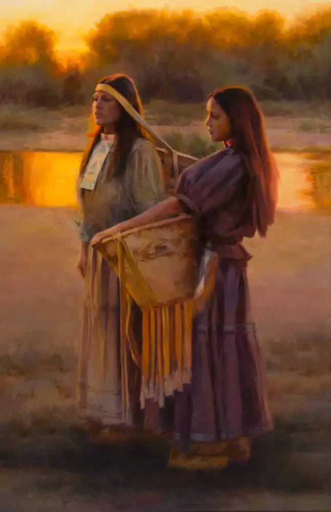 Two women in vintage prairie dresses stand by a sunlit riverbank at sunset, one holding a woven fringed basket.