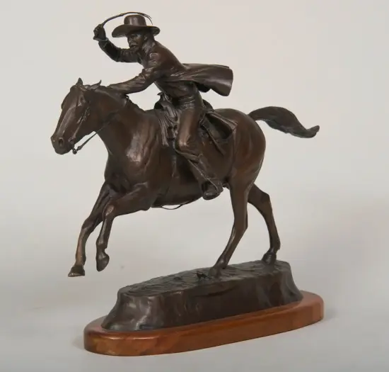 Bronze statue of a horseback rider brandishing a lasso while galloping a rearing horse on a wooden base.