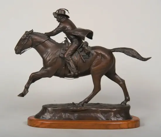 Bronze statue of a cowboy riding a galloping horse, wearing a hat, mounted on a wooden base.