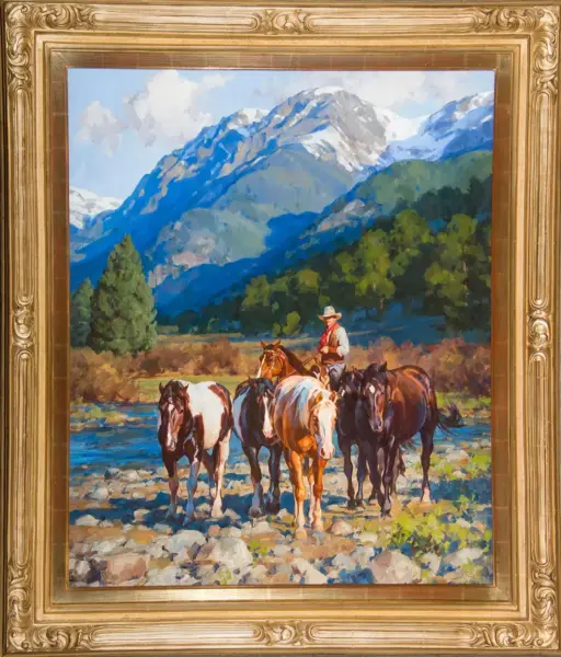 Framed landscape painting of a cowboy on horseback herding several horses across a rocky river with pine trees and snow-capped mountains in the background.
