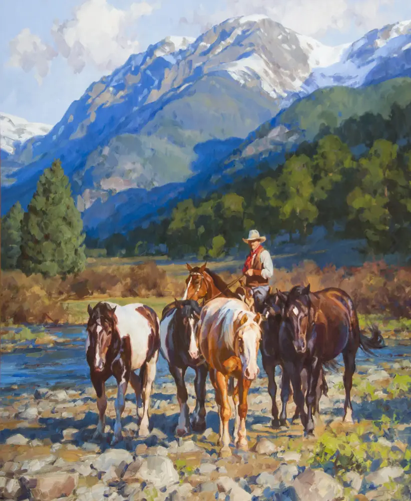 Horses by a river with a rider wearing a hat, set against towering snow-capped mountains in the distance.