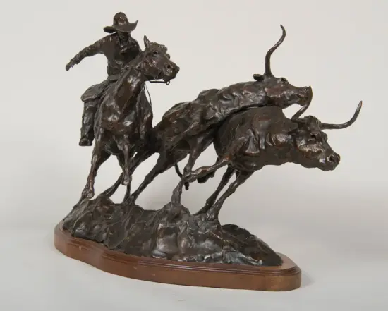 Bronze sculpture of a cowboy on a horse pursuing a charging bull across a rocky base on a wooden pedestal.