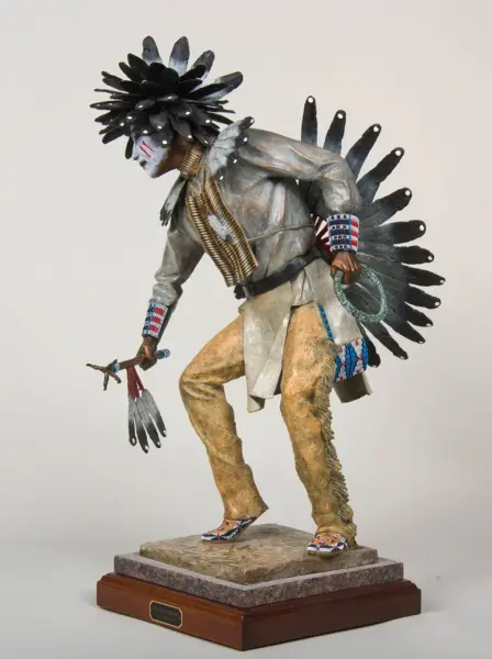 Statue of a Native American man in gray shirt and tan pants with a tall black feather headdress, beaded cuffs, and a feather fan; he stands on a stone pedestal holding a lariat.