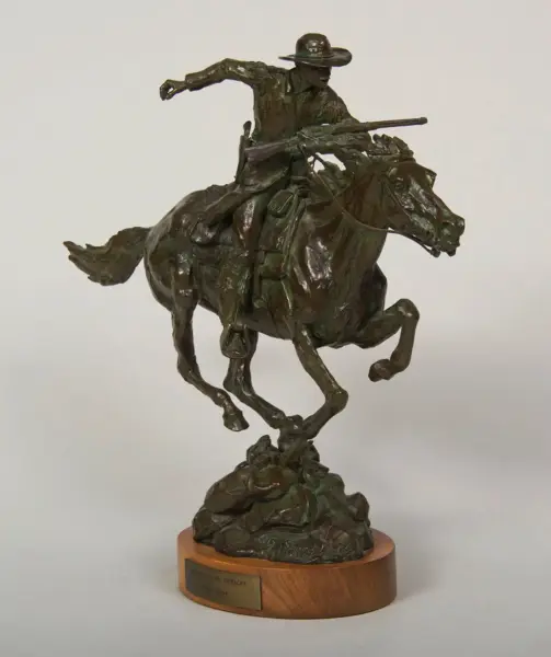 Bronze statue of a cowboy on a rearing horse, aiming a rifle mid-gallop, mounted on a wooden base.