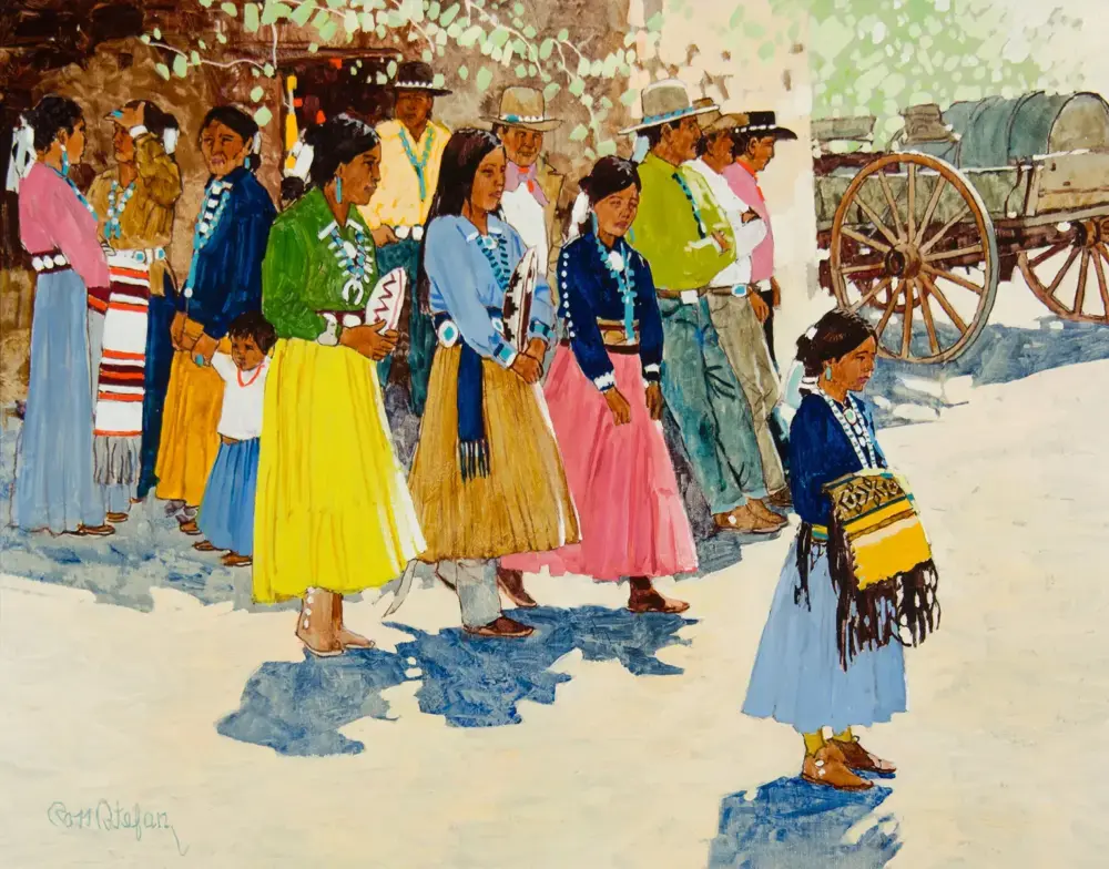 Line of Indigenous women and children in bright traditional dresses and bead necklaces, outdoors near wagon wheels in the background.