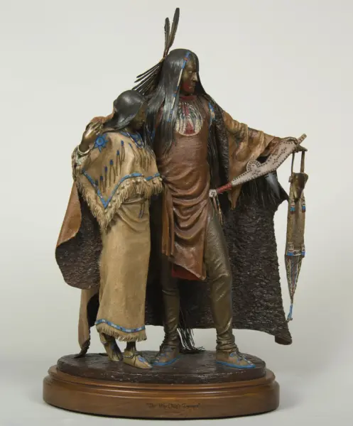 Two Native American figures in traditional dress stand together on a round wooden pedestal; the taller figure wears a feathered headdress and long hair, while the shorter figure rests a hand on the taller's arm. Base reads 'The War Chief's Treasure'.