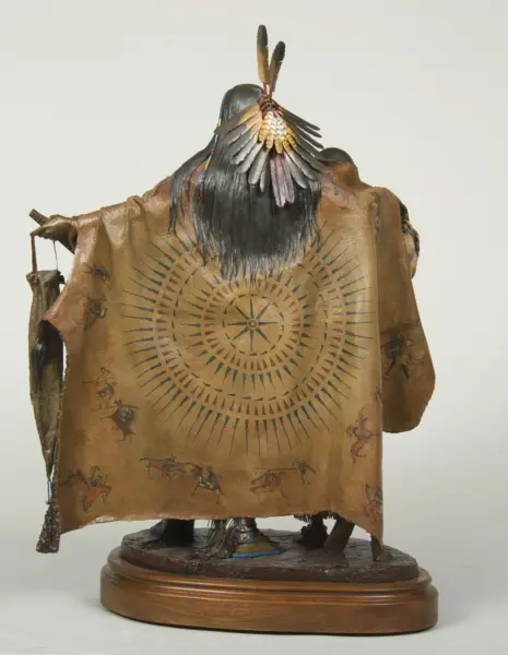 Back view of a native figure in a fringed leather cloak with a sunburst motif and feather headdress on a wooden base.