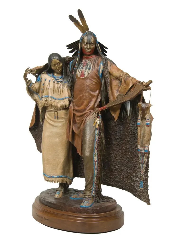 Bronze-toned sculpture of a Native American man in a feathered headdress with a woman beside him, both in fringed beadwork attire on a round pedestal