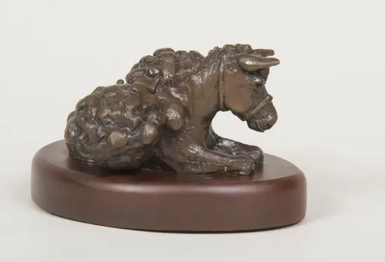 Bronze-toned sculpture of a ram lying down on a circular wooden base, with curled horns and textured wool surface in detail.