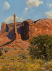 Desert canyon with a tall sandstone spire, red cliffs, and sparse shrub under a blue sky with clouds.