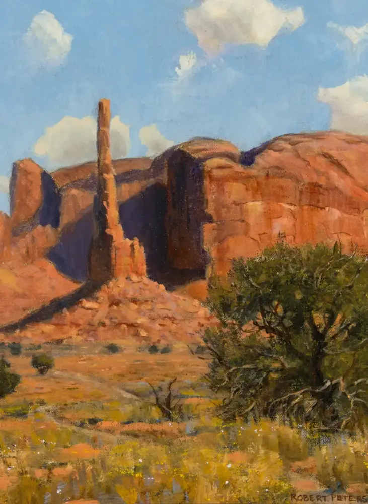 Desert landscape with tall sandstone spire and red rock cliffs under a blue sky with white clouds; desert shrubs in foreground.
