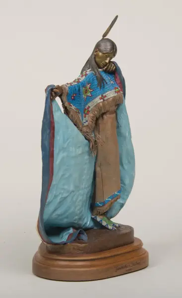 Bronze-toned statue of a Native American woman in a blue beaded shawl, head bowed and holding the edge of her garment, atop a wooden base labeled 'Grandmother's Heart'.