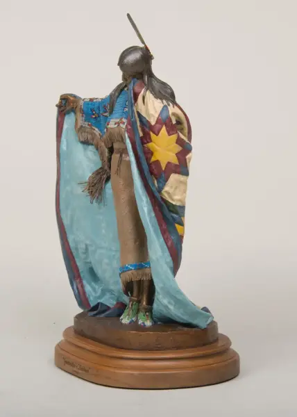 Statue of a Native American man with long hair, wearing a multicolored fringed cloak and quilted blanket, standing on a wooden pedestal.