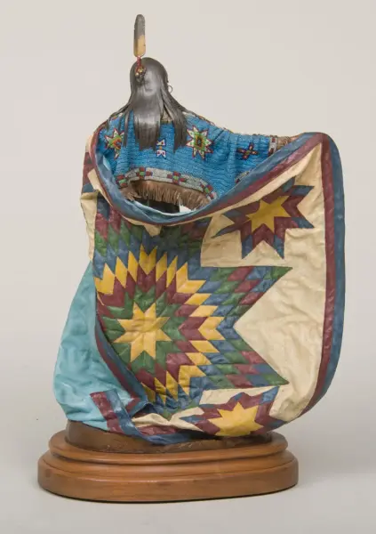 Bust sculpture with long black hair wearing turquoise beadwork and a multicolored star-patterned quilt on a wooden pedestal.