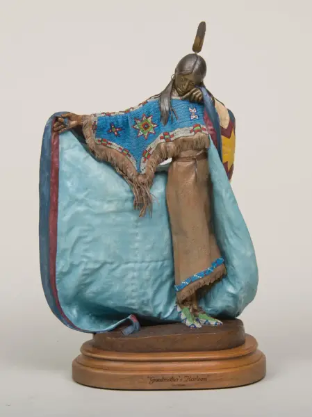 Bronze-toned sculpture of a Native American woman wearing a blue beaded shawl with fringe, standing with arms outstretched on a wooden base labeled “Grandmother’s Heirloom.”