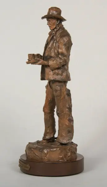 Bronze sculpture of a man in a hat holding a small rectangular object, standing on a textured pedestal (side view).