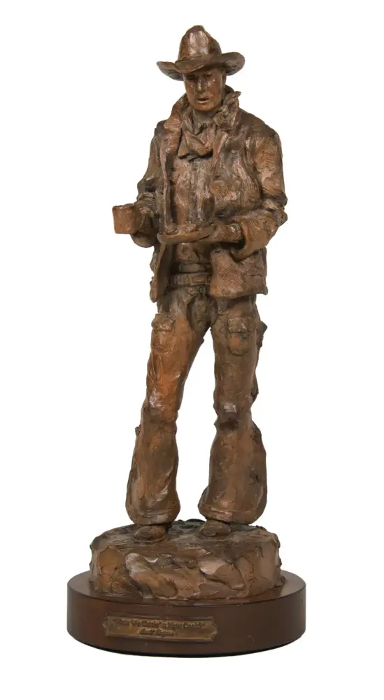 Bronze cowboy statue wearing hat and scarf, holding a mug, standing on a rocky pedestal.