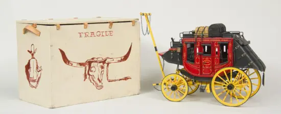Large white crate labeled FRAGILE with a red longhorn drawing, beside a decorative ornate antique horse-drawn carriage on bright yellow wheels.