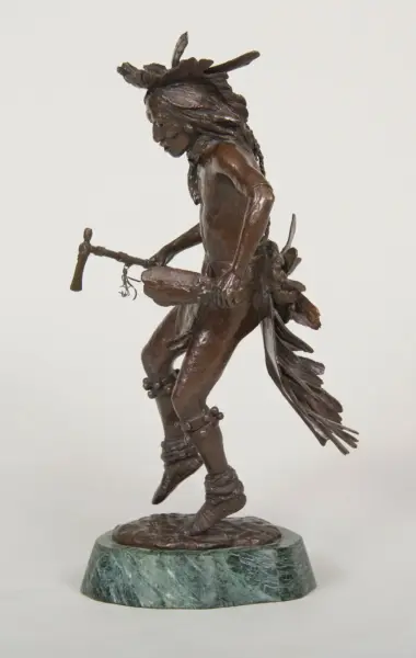Bronze sculpture of a Native American man in traditional dress, with feather headdress, dancing while holding a staff and knife, on a green marble base.