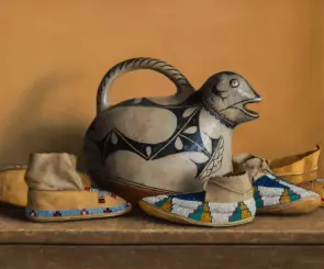 Ceramic bird-shaped pitcher with a curved handle and black geometric designs, set among beaded moccasin footwear on a wooden surface.