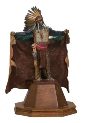 Statue of a Native American chief with a tall feather headdress, outstretched arms, and beaded attire on a wooden pedestal