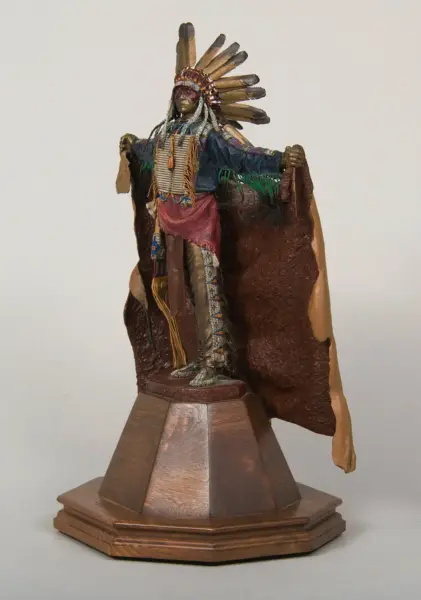 Beaded Native American dancer figurine wearing a feather headdress and fringed garments on a wooden pedestal, posed with arms outstretched.