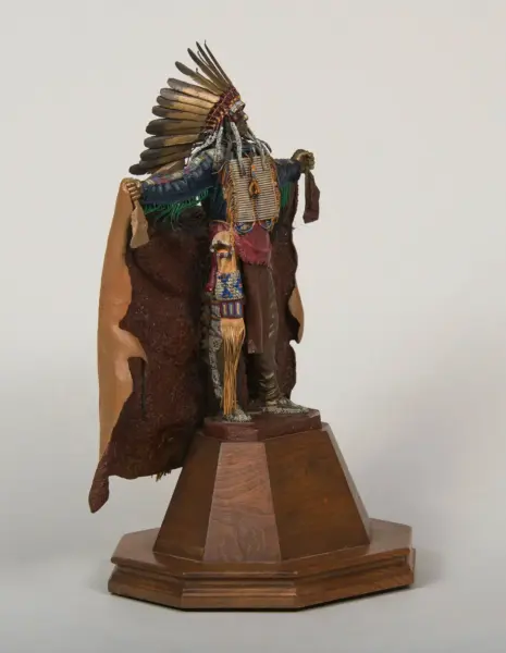 Handcrafted figurine of a Native American man with a large feather headdress, beadwork, and fringed clothing, standing on a rocky base on a wooden pedestal.