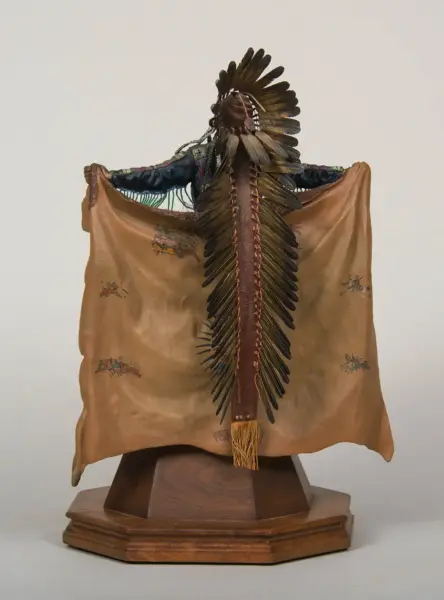 Front-facing display of a feathered headdress and leather cloak on a wooden stand, viewed from the back/side, showcasing long dark feathers and central spine gear swagged with stitches.