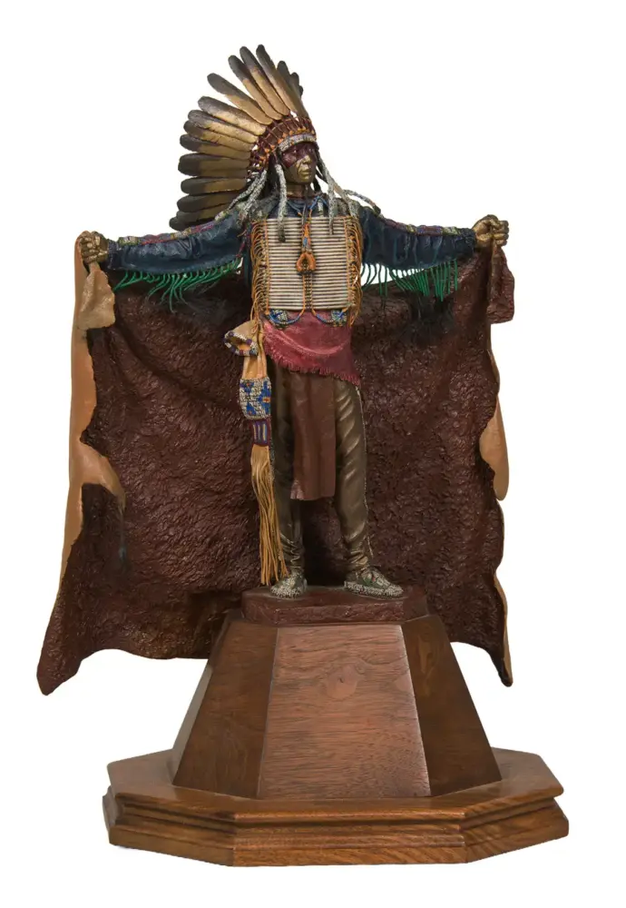 Statue of a Native American chief with a tall feather headdress, outstretched arms, and beaded attire on a wooden pedestal