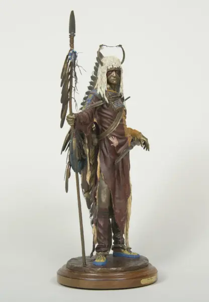 Statue of a Native American warrior in a fringed white headdress, red-brown robes, and jewelry, holding a spear on a wooden base.