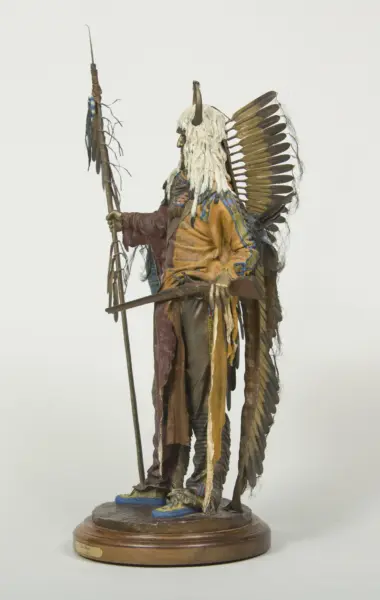 Life-size sculpture of a Native American man in a feathered headdress and fringed regalia, holding a staff, on a wooden pedestal.