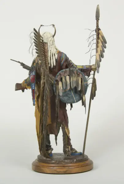 Statue of a humanoid figure in ceremonial dress with feathered wings on the back, holding a tall spear, mounted on a wooden base.