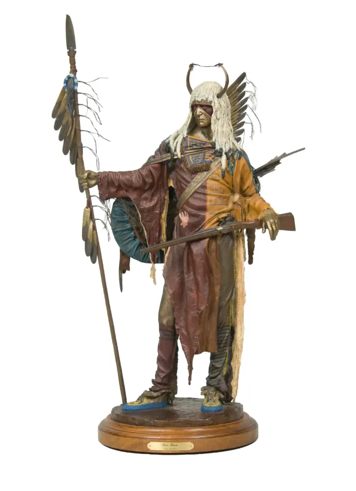 Life-sized statue of a Native American warrior in a feathered headdress, holding a spear and bow on a wooden base.
