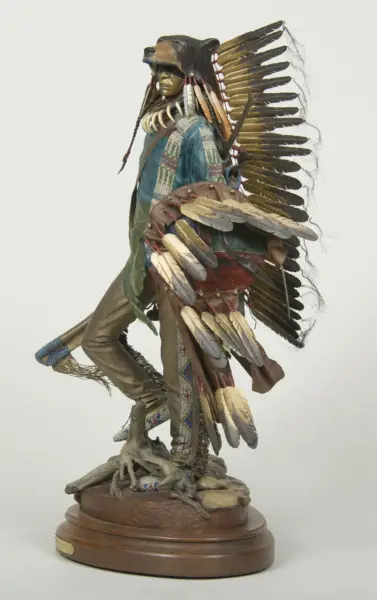 Statue-like figure of a Native American man wearing a large feather war bonnet, beaded clothing, and bone necklace, standing on a rock pedestal