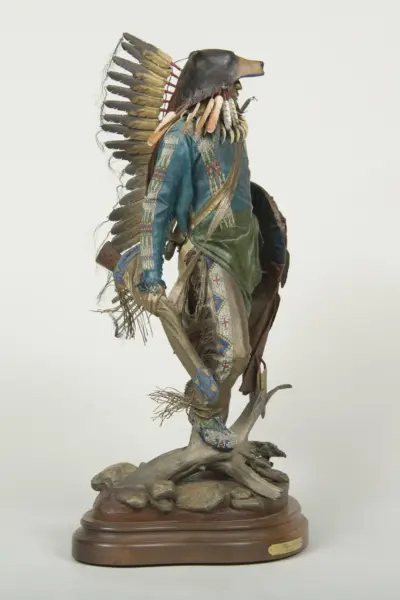 A detailed sculpture of a Native American figure wearing a feathered headdress, beadwork, and fringed clothing on a wooden base