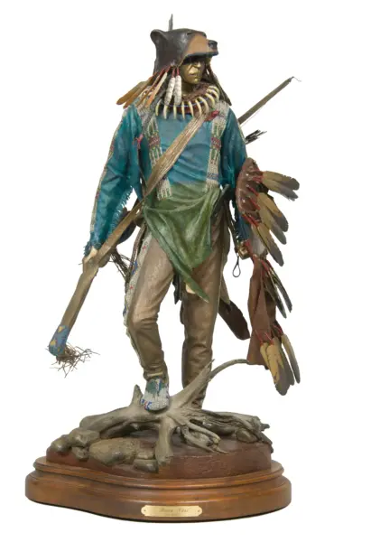 Detailed sculpture of a tribal hunter wearing a bear head headdress, turquoise robes, and bone necklace, standing on a rocky wooden base.