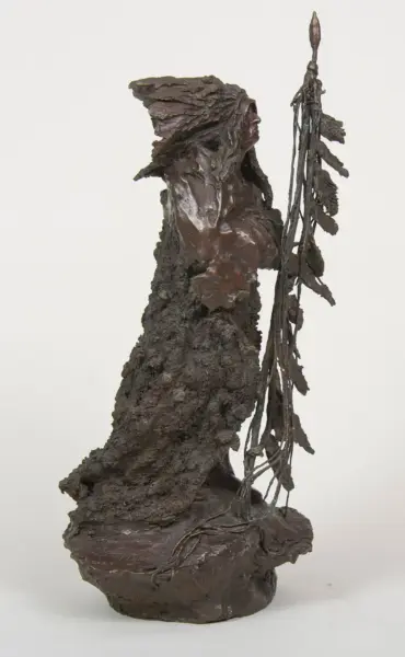 Bronze sculpture of a robed, elongated figure holding a tall staff wrapped in vines, viewed from the side against a light background.