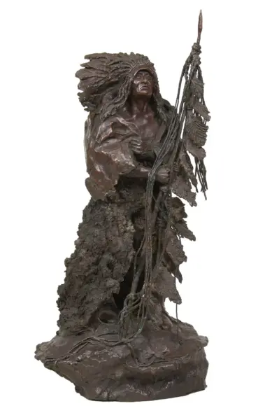 Bronze sculpture of a Native American man with a feathered headdress holding a tall staff on a rocky base.