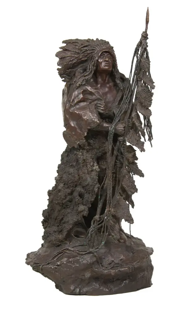 Bronze sculpture of a Native American man with a feathered headdress holding a tall staff on a rocky base.