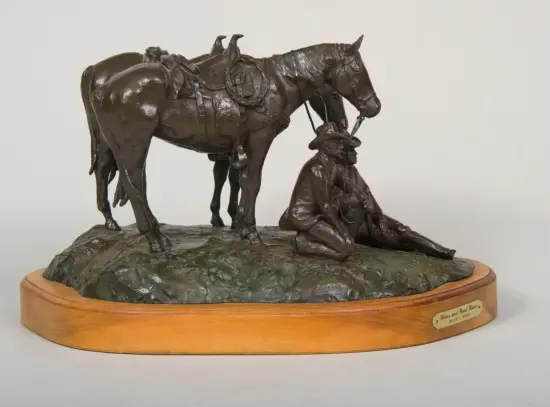 Bronze sculpture of a cowboy kneeling beside a saddled horse on a rocky base, mounted on a wooden pedestal plaque.