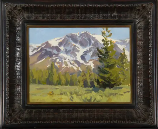 Oil painting of a snow-capped mountain range with evergreen trees in a sunlit meadow, framed in a dark carved frame.