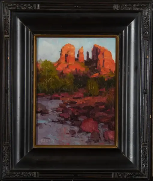 Oil painting of red rock formations and green trees in a desert landscape, inside a heavy black carved frame.