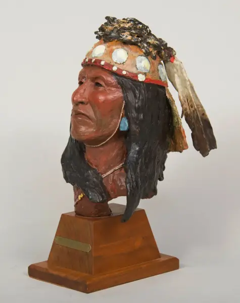 Bust sculpture of a Native American man in a feathered headdress on a wooden pedestal.